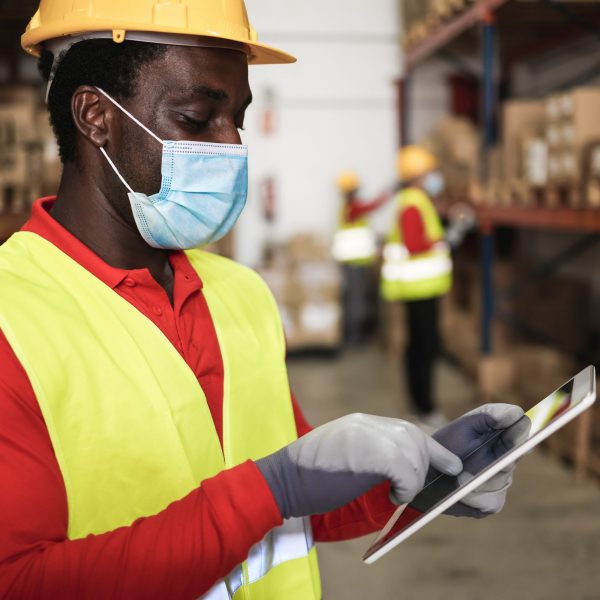 African worker man using tablet inside warehouse while wearing safety mask - Focus on face